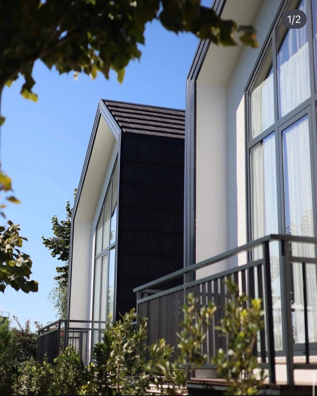 A picturesque corner house with traditional architectural style featuring wooden accents and lush green ivy climbing up the walls. The building has multiple windows adorned with red flower boxes, and the ground floor features a sign indicating it's a hotel. The house is painted a light green color, and there are cobblestone streets surrounding it.